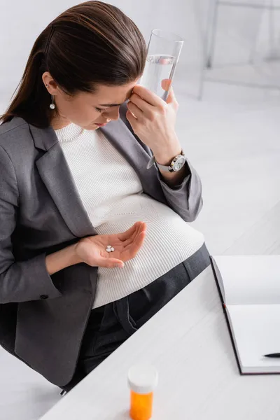 Беременная женщина держит таблетки и стакан воды, чувствуя спазм в офисе — стоковое фото