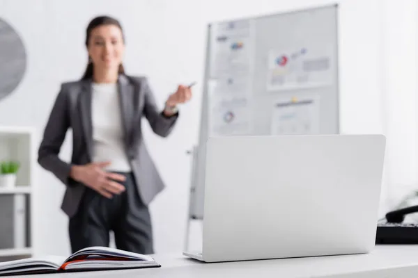 Laptop nahe schwangerer Frau nahe Flipchart auf verschwommenem Hintergrund — Stockfoto