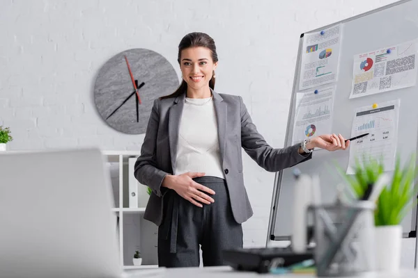 Cheerful pregnant businesswoman pointing at flip chart during online conference — Stock Photo
