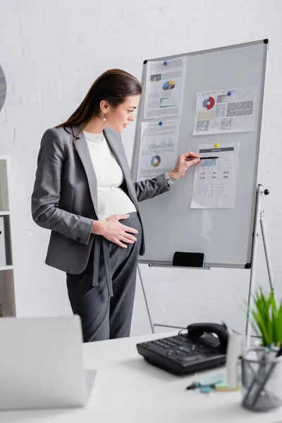 Donna incinta indicando lavagna a fogli mobili vicino al computer portatile offuscato sulla scrivania — Foto stock