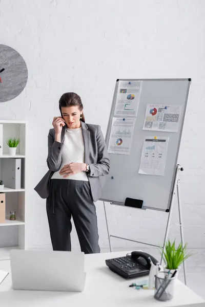 Focused pregnant woman looking at laptop on desk while standing near flip chart — Stock Photo