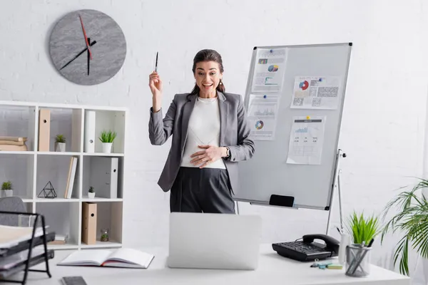 Mujer embarazada emocionada mirando a la computadora portátil en el escritorio mientras está de pie cerca del rotafolio - foto de stock