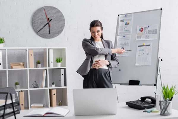 Fröhliche schwangere Geschäftsfrau zeigt auf Flipchart während Online-Konferenz auf Laptop — Stockfoto