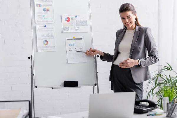 Cheerful pregnant businesswoman pointing at flip chart with chats and graphs during online conference on laptop — Stock Photo