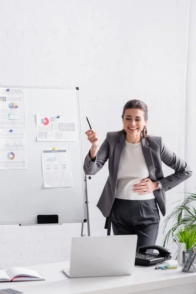 Jovem mulher de negócios grávida rindo perto de flip chart com gráficos durante chamada de vídeo no laptop — Fotografia de Stock