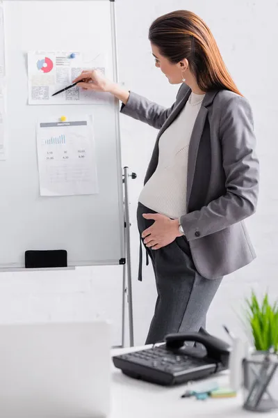 Fröhliche schwangere Geschäftsfrau zeigt mit Stift auf Flipchart — Stockfoto