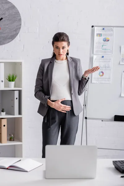 Joven embarazada mujer de negocios gesto cerca de rotafolio con chats y gráficos durante la videollamada en el ordenador portátil - foto de stock