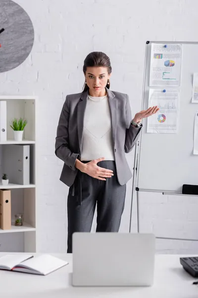 Jeune femme d'affaires enceinte gestuelle près de flip chart avec des chats et des graphiques lors d'un appel vidéo sur ordinateur portable — Photo de stock