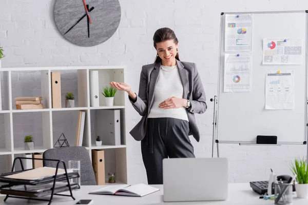 Giovane donna d'affari incinta gesticolando vicino flip chart con grafici durante la videochiamata sul computer portatile — Foto stock