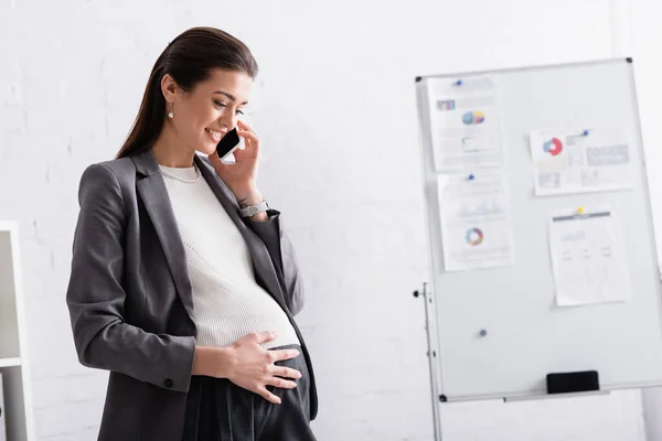 Alegre embarazada mujer de negocios hablando en smartphone cerca de rotafolio en la oficina - foto de stock