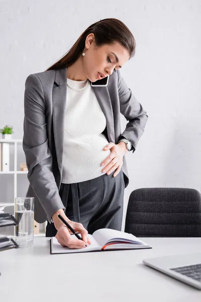 Empresaria embarazada escribiendo en cuaderno y hablando por celular - foto de stock