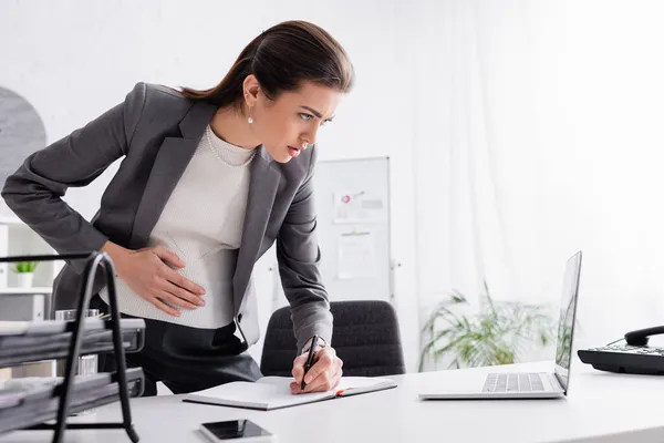 Focused pregnant businesswoman writing in notebook near devices on table — Stock Photo