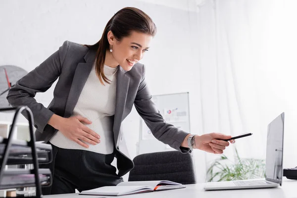 Feliz embarazada mujer de negocios señalando con pluma en el ordenador portátil en el escritorio - foto de stock