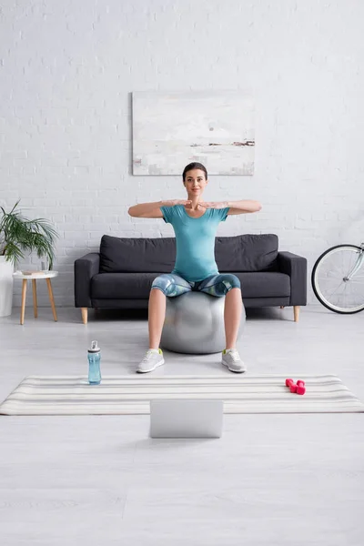 Happy pregnant woman in sportswear exercising on fitness ball near laptop in living room — Stock Photo