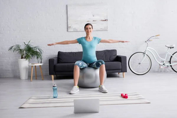 Mujer embarazada alegre en ropa deportiva ejercicio en la pelota de fitness cerca de la computadora portátil en la sala de estar - foto de stock