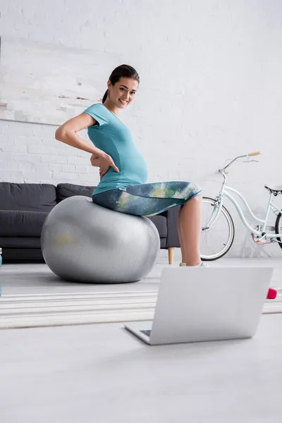 Позитивна вагітна жінка в спортивному одязі вправляється на фітнес-кулі і дивиться на ноутбук у вітальні — стокове фото