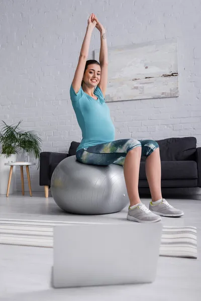 Positive pregnant woman with raised hands exercising on fitness ball and looking at laptop in living room — Stock Photo