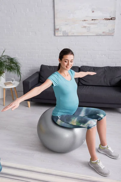 Улыбающаяся беременная женщина с протянутыми руками, упражняющаяся на фитнес-мяч в гостиной — стоковое фото
