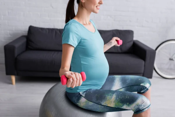 Cropped view of smiling pregnant woman exercising on fitness ball with pink dumbbells — Stock Photo