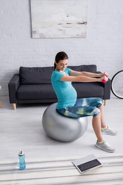 Sonriendo mujer embarazada haciendo ejercicio en la pelota de fitness con pesas cerca de la computadora portátil - foto de stock