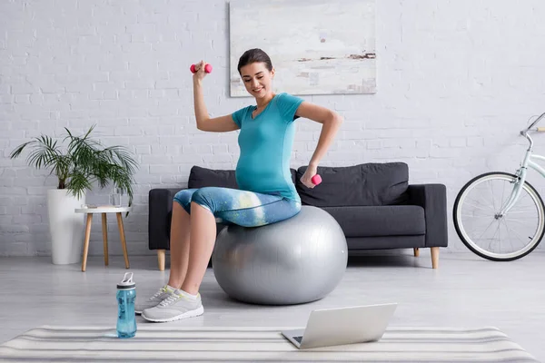 Mujer embarazada alegre haciendo ejercicio en la pelota de fitness con pesas cerca de la computadora portátil - foto de stock