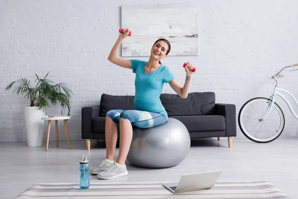 Mulher grávida feliz exercitando na bola de fitness com halteres perto do laptop — Fotografia de Stock