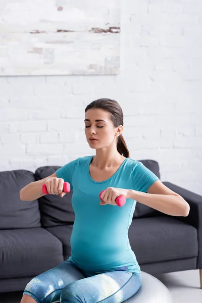 Pregnant sportswoman exercising on fitness ball with pink dumbbells — Stock Photo