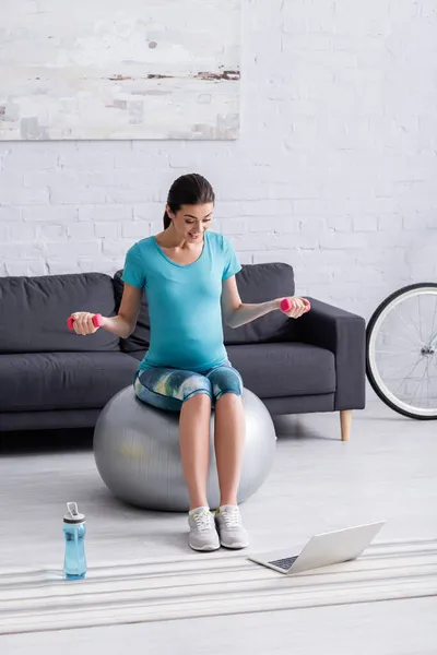 Joyful pregnant sportswoman exercising on fitness ball with dumbbells near sports bottle and laptop — Stock Photo