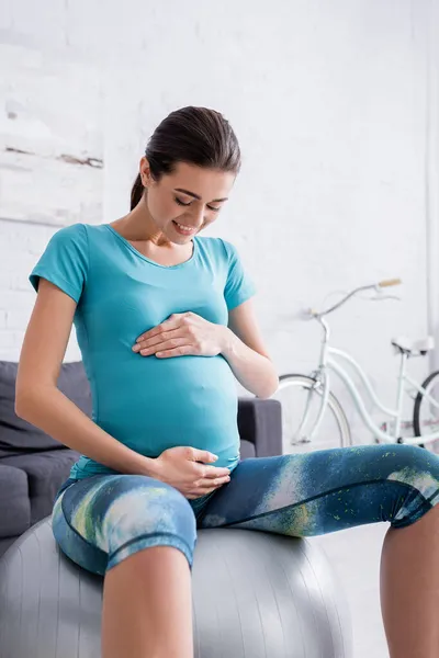 Alegre embarazada deportista sentado en la pelota de fitness y tocando el vientre - foto de stock