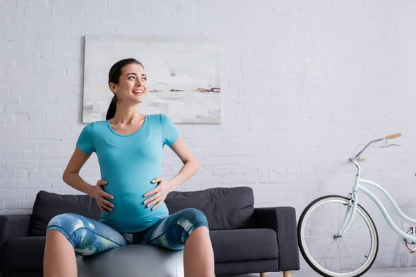 Deportista embarazada alegre sentado en la pelota de fitness y mirando hacia otro lado - foto de stock