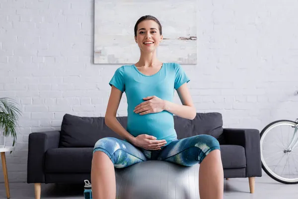 Радостная беременная спортсменка сидит на фитнес-мяч и смотрит в камеру — стоковое фото