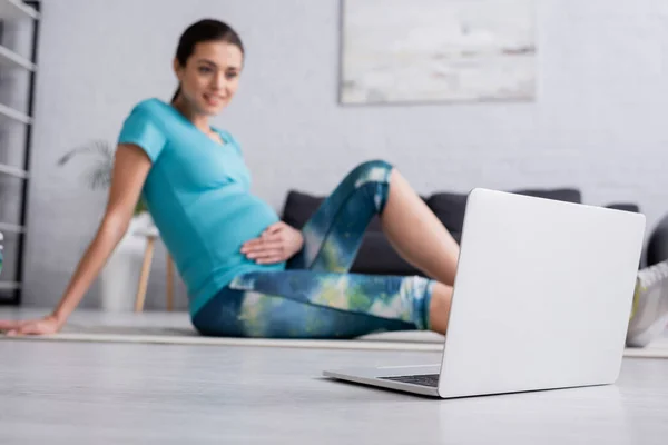 Весела вагітна жінка в спортивному одязі, сидячи на фітнес-маті біля ноутбука у вітальні — стокове фото