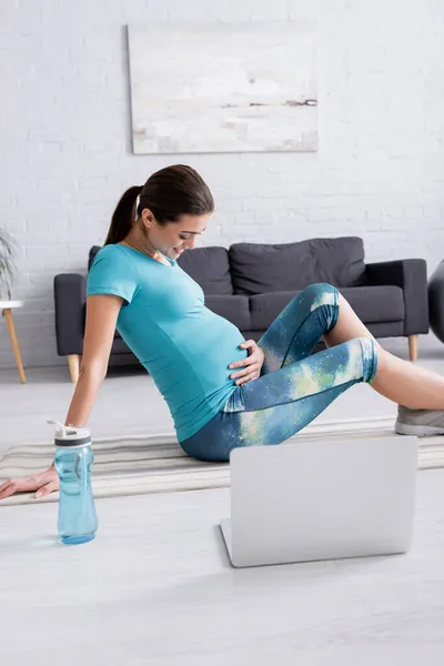 Беременная женщина в спортивной одежде сидит на тренажерном коврике и смотрит на живот возле ноутбука в гостиной — стоковое фото