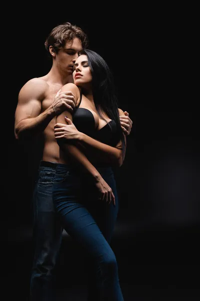 Без сорочки чоловік в джинсах обіймає брюнетку жінку в бюстгальтері на чорному — стокове фото