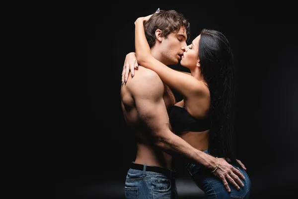 Vue latérale de jeune et sexy couple en jeans baisers sur noir — Photo de stock