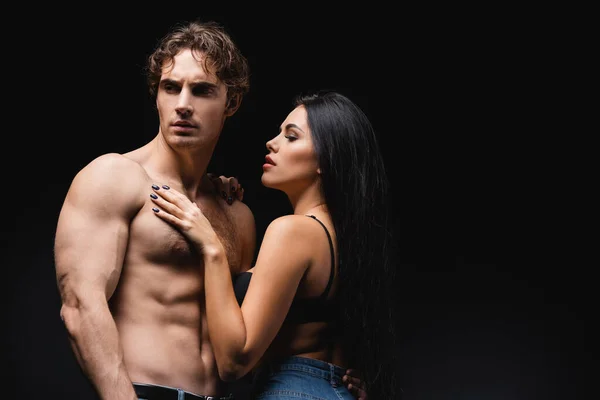 Young woman in bra and jeans touching muscular boyfriend isolated on black — Stock Photo