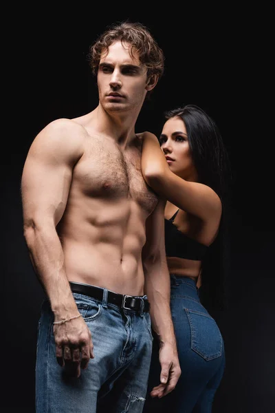 Junge Frau in BH und Jeans stützt sich auf muskulösen Freund isoliert auf schwarz — Stockfoto