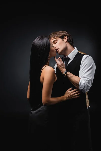 Homem em colete e camisa beijando namorada em vestido de deslizamento sexy no preto — Stock Photo