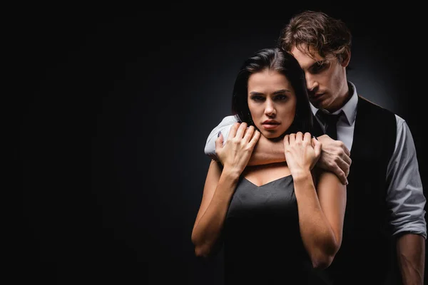Mann in Weste und Hemd umarmt und erstickt sexy Freundin in Seidenkleid isoliert auf schwarz — Stockfoto