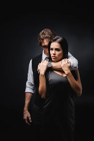 Мужчина в жилете и белой рубашке обнимает и душит сексуальную девушку в шелковом платье на черном — стоковое фото