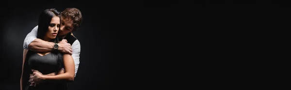 Homme en gilet et chemise blanche étreignant petite amie brune en soie robe glissante sur noir, bannière — Photo de stock