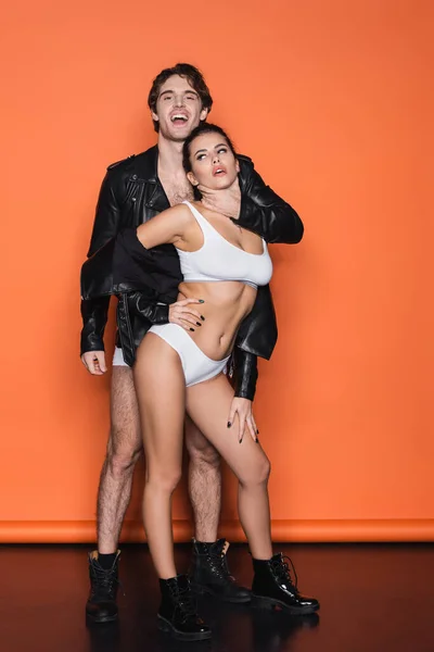 Piena lunghezza di uomo felice in giacca di pelle nera soffocamento donna sexy in lingerie su arancione — Foto stock
