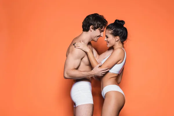 Vue latérale du couple heureux et sexy en sous-vêtements blancs debout sur orange — Photo de stock