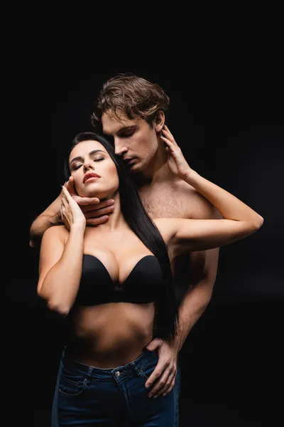 Sexy young woman in bra and jeans embracing muscular man isolated on black — Stock Photo