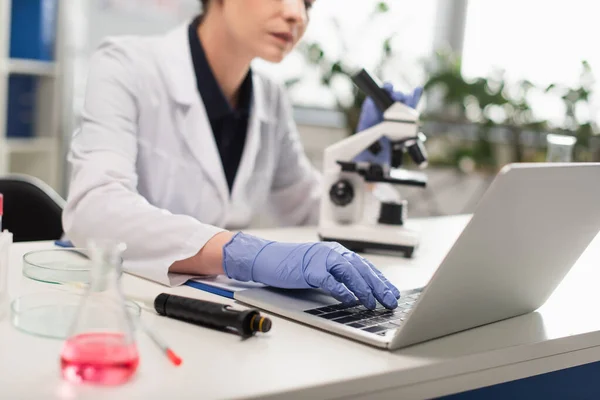 Cropped view of blurred scientist in latex gloves using laptop near equipment — Stock Photo