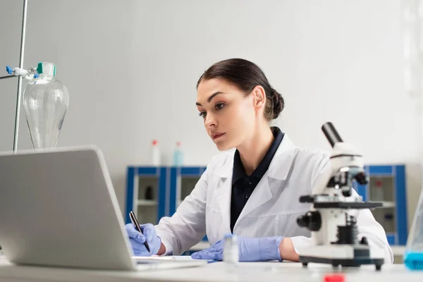 Científico en guantes de látex escribir en portapapeles cerca de la computadora portátil y microscopio en el laboratorio - foto de stock