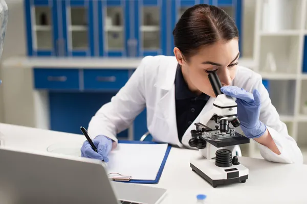 Scientifique regardant le microscope près du presse-papiers et de l'ordinateur portable en laboratoire — Photo de stock