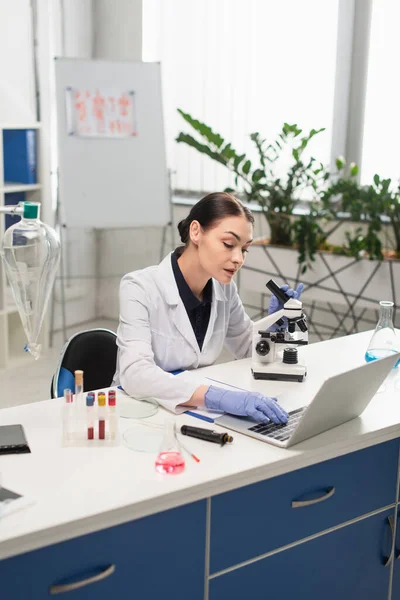 Científico morena utilizando portátil cerca del microscopio y el equipo en el laboratorio - foto de stock