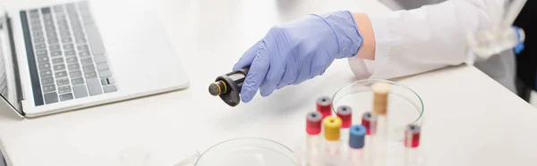 Cropped view of scientist in latex glove holding electronic pipette near laptop and test tubes in lab, banner — Stock Photo