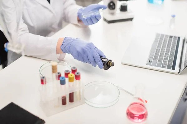 Vista recortada del científico en guantes de látex que sostiene la pipeta electrónica cerca de la computadora portátil y tubos de ensayo - foto de stock